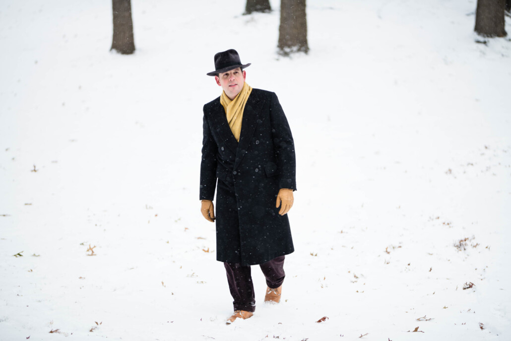 A photograph of Raphael walking in the snow while wearing a dark overcoat and yellow accessories.
