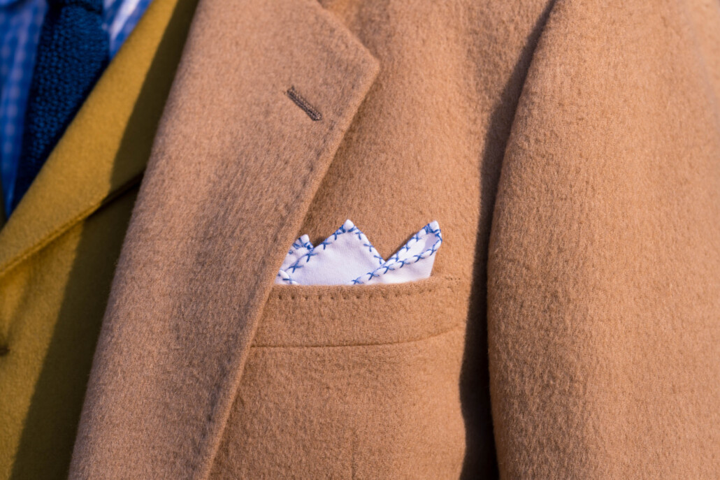 A white pocket square with blue edging in a camel hair coat pocket