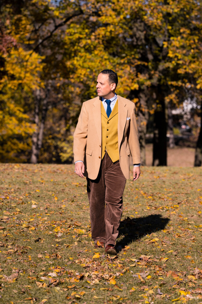 Raphael walks through a fall scene in a camel jacket with brown trousers and yellow waistcoat