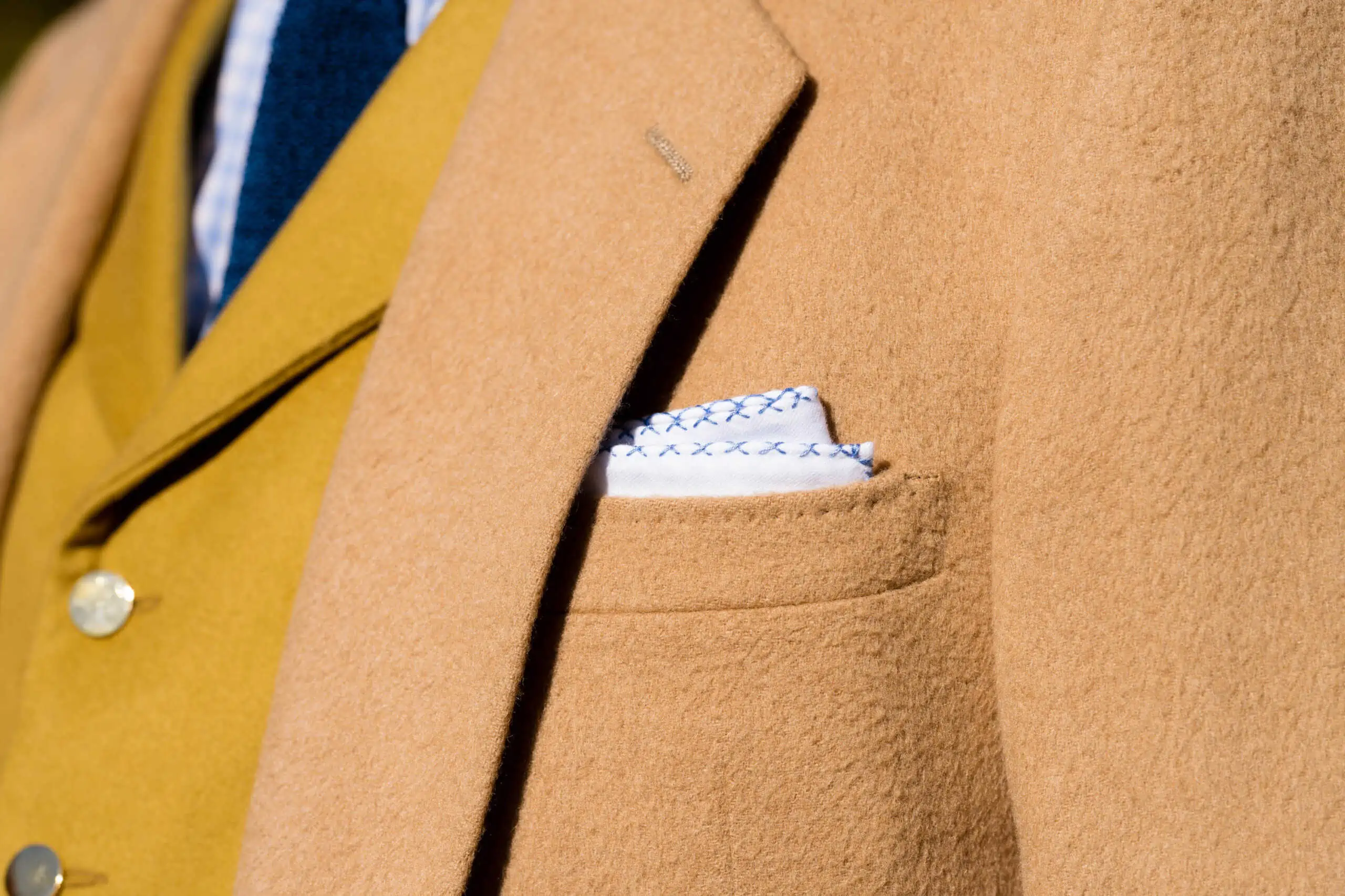 A white pocket square with blue edging folded in a TV or banker square in a camel hair jacket pocket