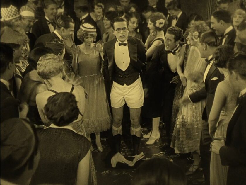 Still from a film showing a man standing in his underwear at a party