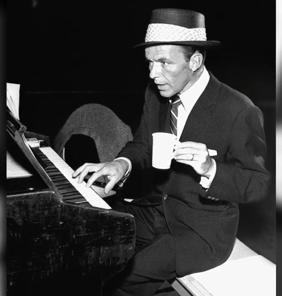 Photo of Frank Sinatra playing piano with coffee mug dark suit repp tie bold hat band