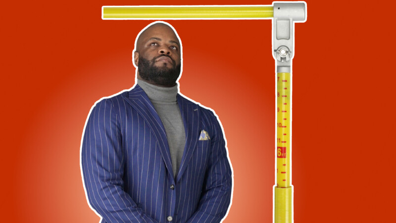 Kyle, in a blue pinstriped suit and gray turtleneck sweater, looks upward at a height measurement bar.