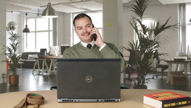 David Sperber, in a green v-neck sweater, white shirt, and brown tie, sits at a desk while talking on the phone, as a laptop, tie, and business book sit in front of him, with an office scene behind