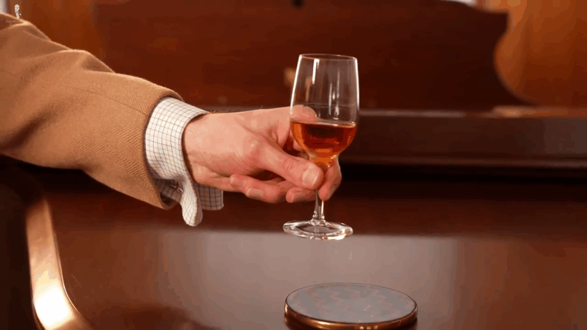 Hand pinching the mid-stem of a small fortified wine glass, holding it delicately with two fingers.