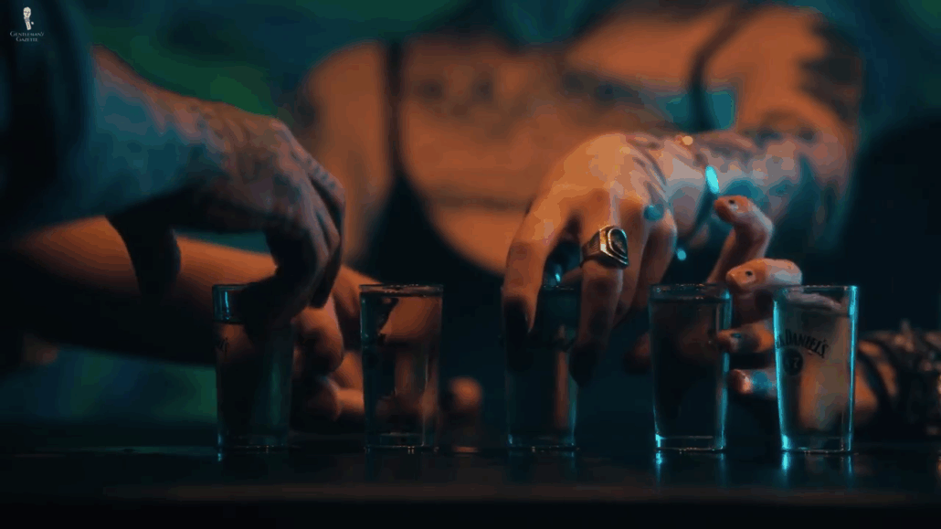  Hands lightly pinching a shot glass at the base, avoiding wrapping the hand around it.