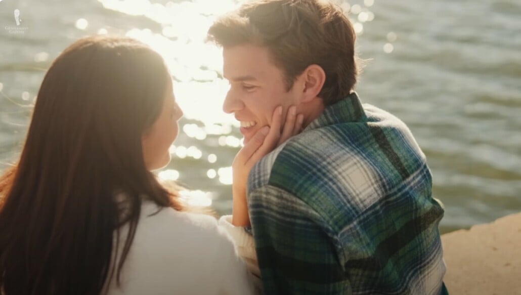 Gentleman Vice Philandering Young couple sitting by the water at sunset, smiling as the woman gently touches the man’s face while they gaze at each other affectionately
