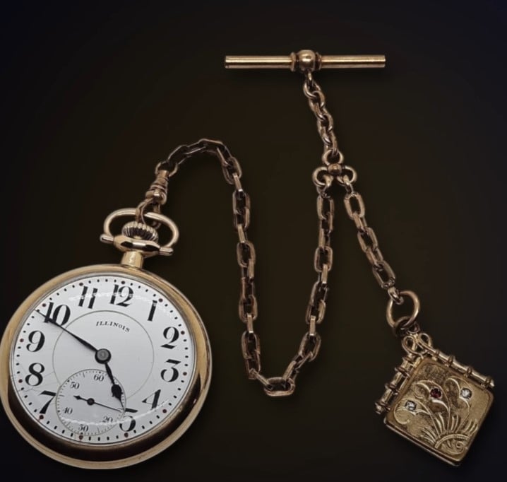 gold illinois pocket watch decorative fob A gold-toned Illinois pocket watch with a white face, black numerals, and a subsidiary seconds dial at the 6 o'clock position. It is attached to a matching chain featuring a T-bar and an ornate, rectangular fob with floral engravings and small gemstones.