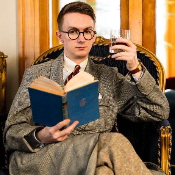 ome attire mens classic lounge style Man in a plaid robe and tie sitting in an ornate armchair, holding a glass of water and reading a blue hardcover book, with playing cards on a side table in the foreground.
