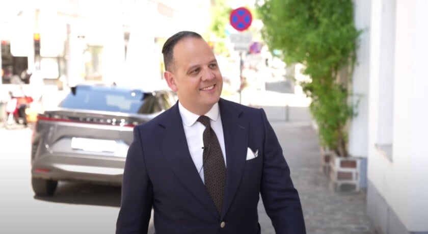 raphael outfit rundown hedi part two vienna Raphael in a navy two-button suit with a white shirt, dark brown textured tie, and a white linen pocket square, smiling while walking down a sunlit Vienna street.