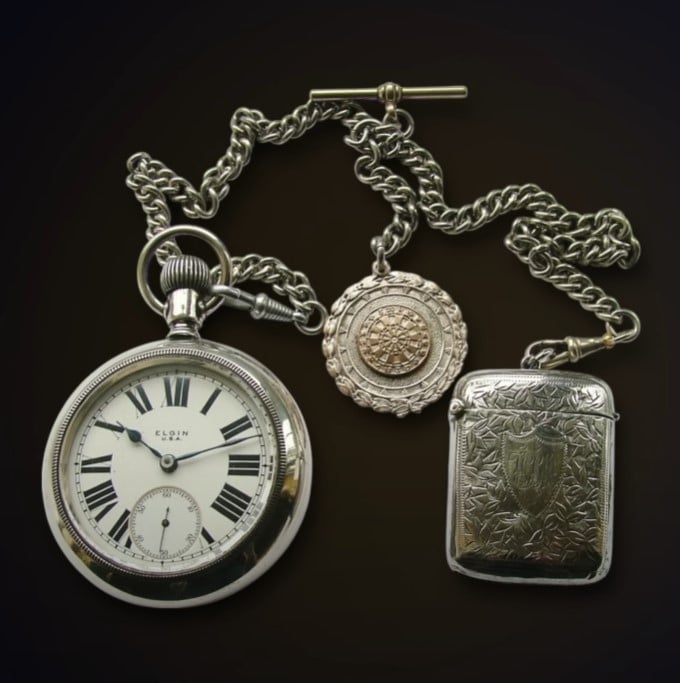 silver elgin pocket watch multiple fobs A silver Elgin pocket watch with a white face, black Roman numerals, and a subsidiary seconds dial. It is connected to a silver chain with a T-bar, a circular, multi-layered fob with gold accents, and a rectangular, engraved match safe or vesta case.