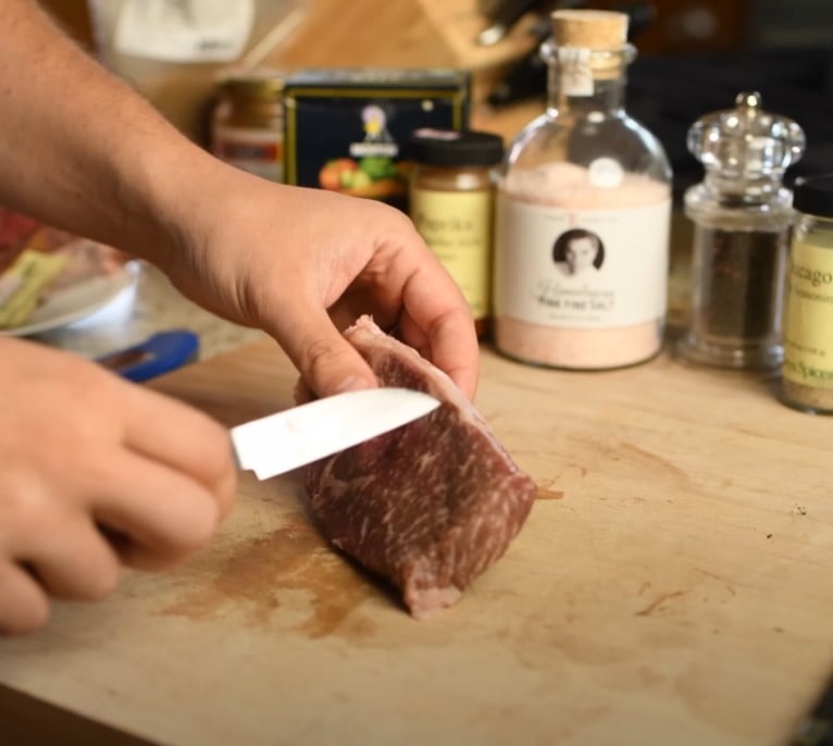 slicing wagyu beef fine dining Person slicing marbled wagyu beef on a wooden cutting board with spices in the background.