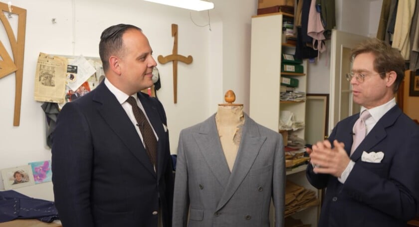 Raphael Outfit Mass Salon Possanner Visit Raphael Schneider, in a navy suit and brown tie, listens as tailor Michael Possanner explains his craft in his Viennese workshop, with a grey jacket on a mannequin between them.