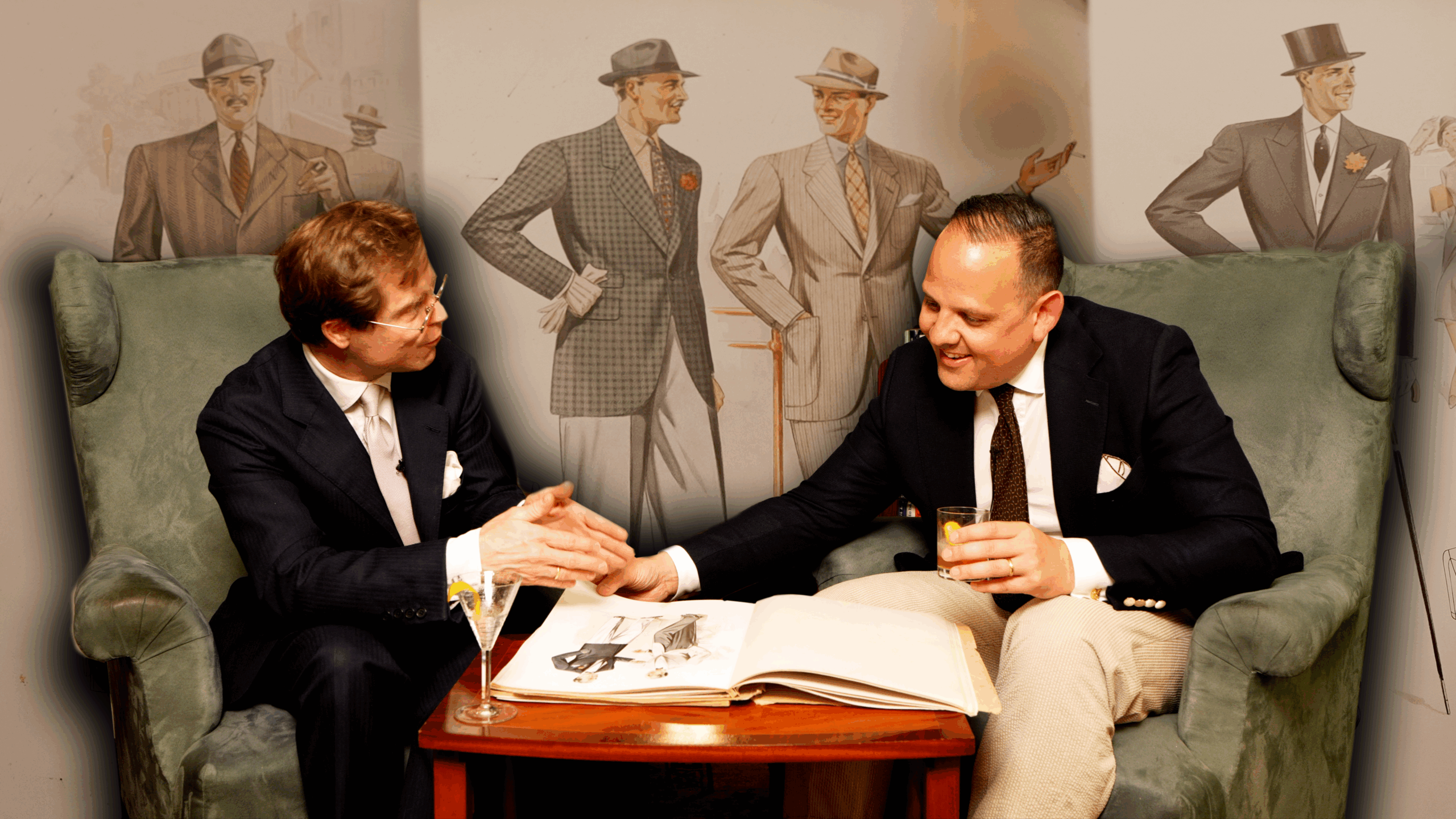 Two men in formal attire sit in green wingback chairs, gesturing toward an open book of fashion plates on a mahogany table. The background&mdash;a wall adorned with large-scale 1940s-style men's fashion illustrations&mdash;is rendered with a soft blur to create depth.