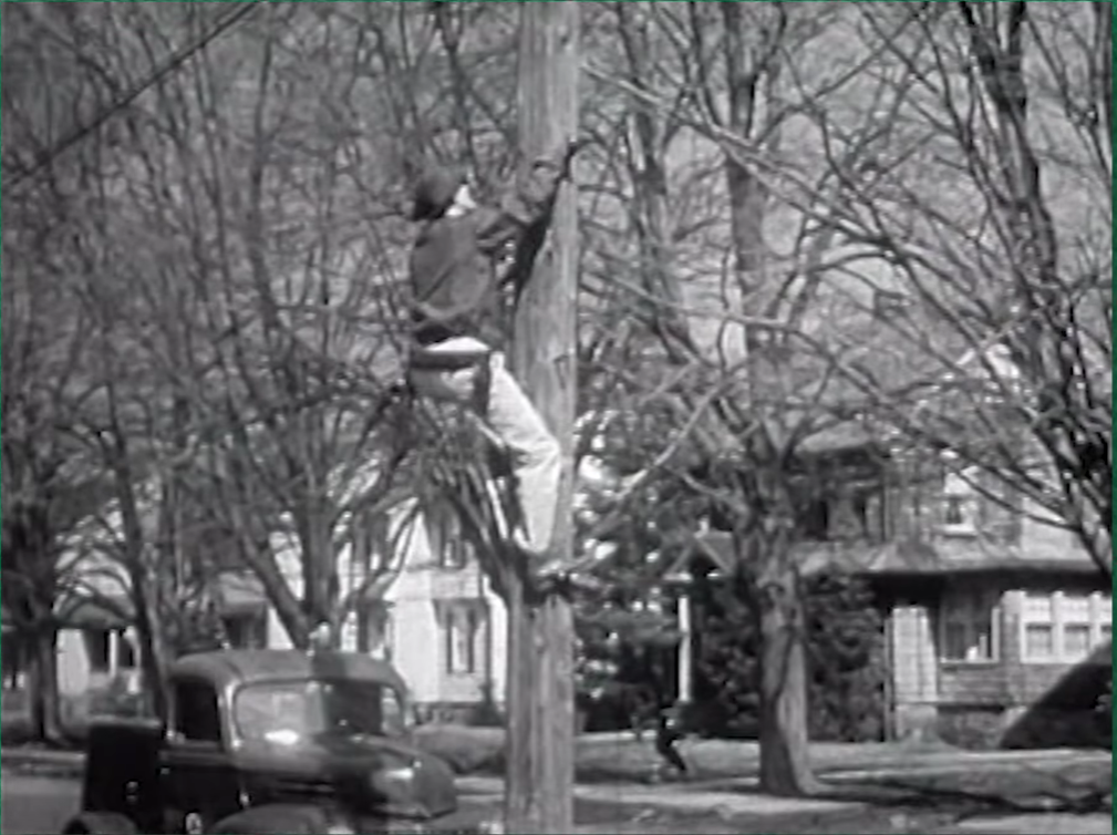 A vintage black and white photo of a utility worker climbing a wooden pole.