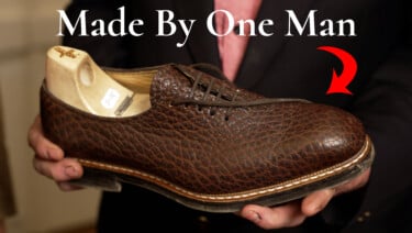Handmade brown textured leather dress shoe with a wooden shoe tree being held by a craftsman; text reads: “Made By One Man.”