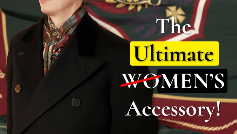 A man in a dark double-breasted overcoat wears a patterned silk scarf tied at the neck, standing in front of a large decorative scarf backdrop. Text reads: &ldquo;The Ultimate Women&rsquo;s Accessory!&rdquo; with &ldquo;Women&rsquo;s&rdquo; crossed out in red.