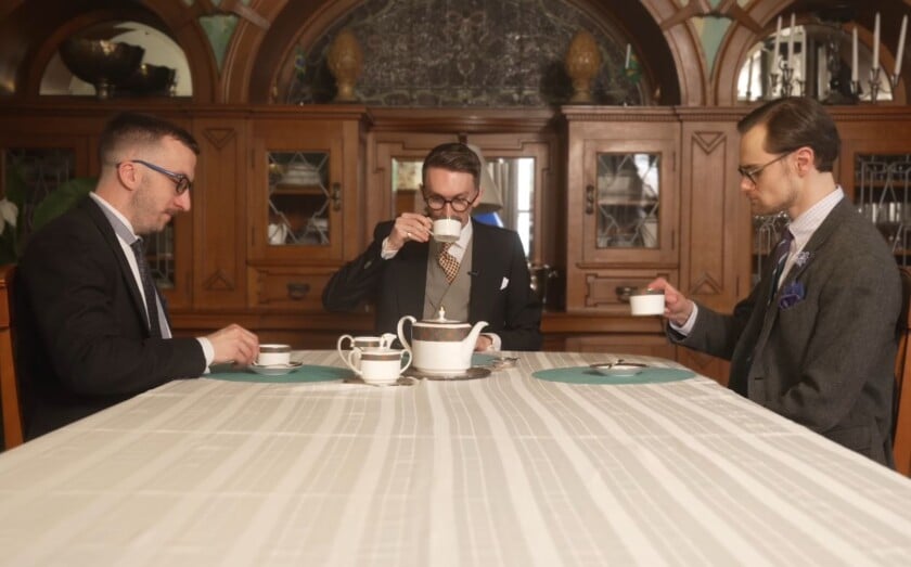 Three men seated at a table drinking tea, lifting only the teacup while the saucer remains on the table.