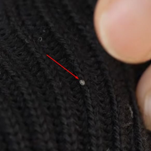 Macro close-up of a single, tiny, white, rice-shaped clothes moth egg nestled in the ribs of a black wool knit garment, highlighted by a red arrow.