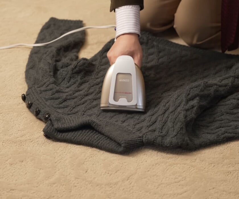 Close-up of a person using a handheld Conair garment steamer on a grey cable-knit wool sweater to kill clothes moth larvae through heat treatment.