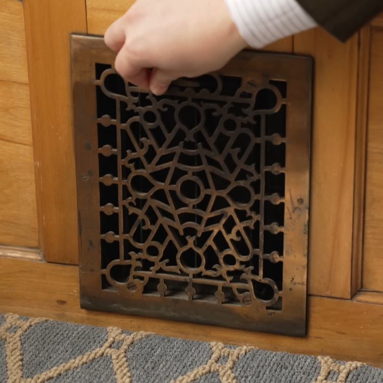 A hand lifting a metal floor register to inspect the dusty ductwork underneath for moth activity.
