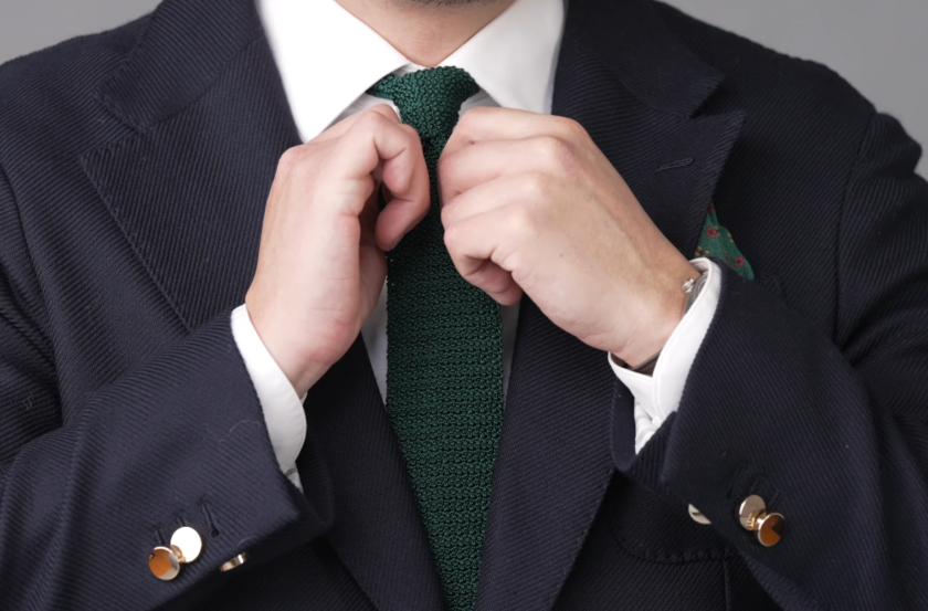 Ivan adjusting a dark green silk knit tie worn with a white shirt and navy textured jacket with gold buttons.