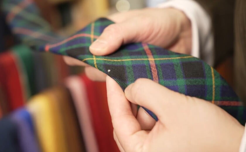 Close-up of a small, white hole on the edge of a green and blue tartan wool tie, where clothes moth larvae have eaten through the wool fabric to reveal the inner lining.