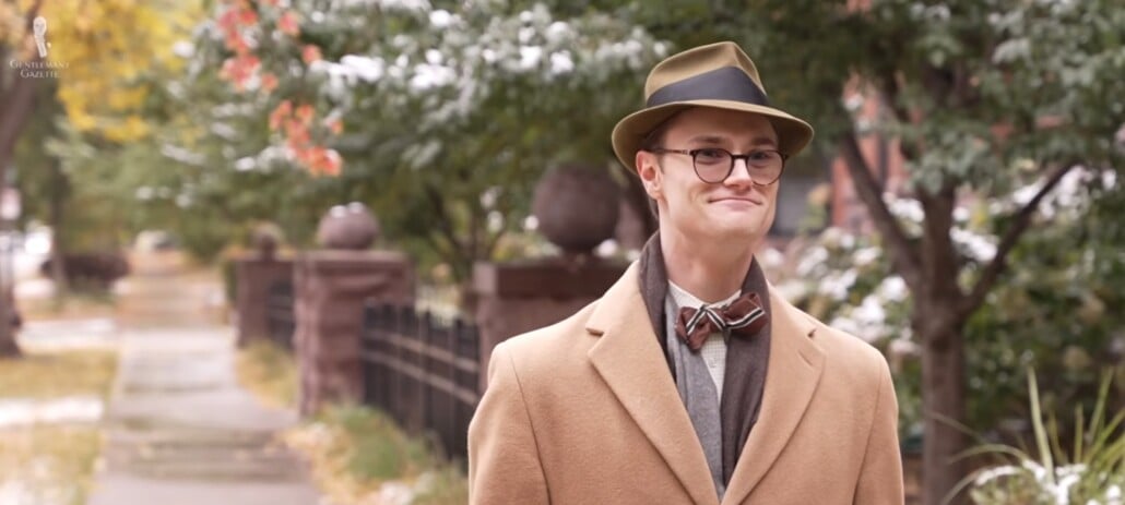 Preston walks down a street wearing a camel hair coat and matching hat.