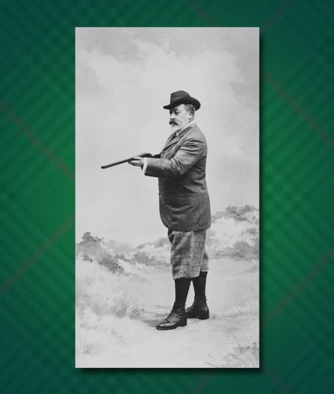 A vintage black-and-white full-length photograph of King Edward VII standing in a field during a hunting expedition. He is shown in profile, wearing a Homburg hat, a patterned tweed jacket, and knee-length knickerbockers with tall socks and lace-up boots