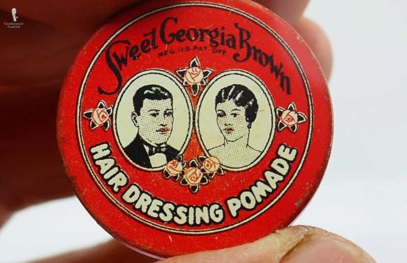 Vintage Sweet Georgia Brown Hair Pomade Tin A close-up of a small, vintage red tin of Sweet Georgia Brown Hair Dressing Pomade being held by two fingers