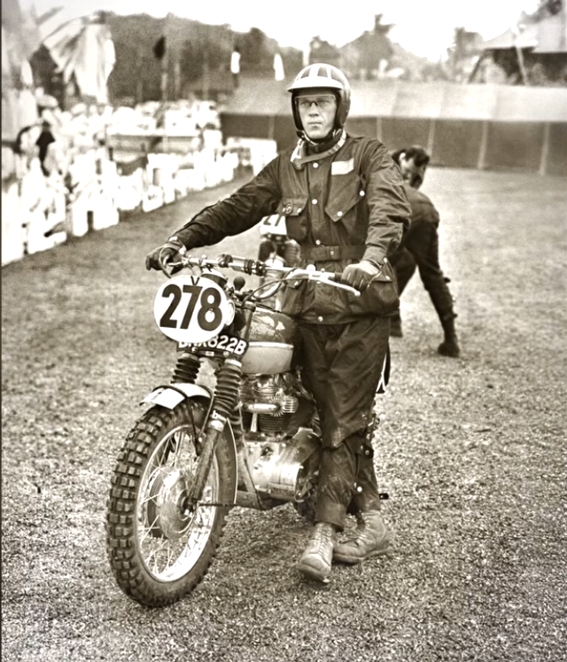 Black and white photo of a motorcycle racer in full racing suit, helmet, and glasses, standing beside a competition motorcycle numbered 278 at a race event