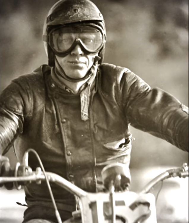 Sepia-toned close-up of a motorcycle racer in a worn leather jacket, helmet, and goggles, leaning forward on a racing motorcycle with number 163