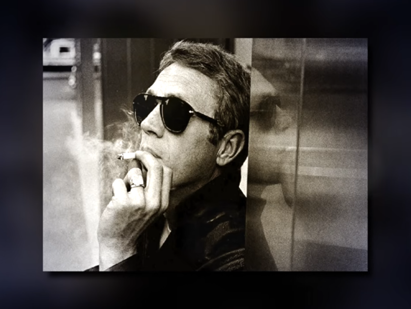 Black and white photo of Steve McQueen smoking a cigarette, wearing dark sunglasses and a signet ring, his reflection visible in glass behind him