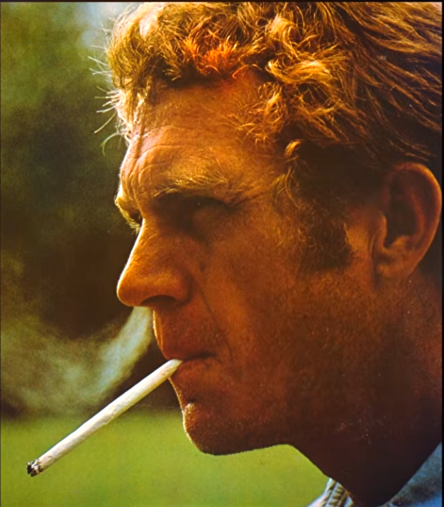 Close-up color profile shot of Steve McQueen smoking an unfiltered cigarette outdoors