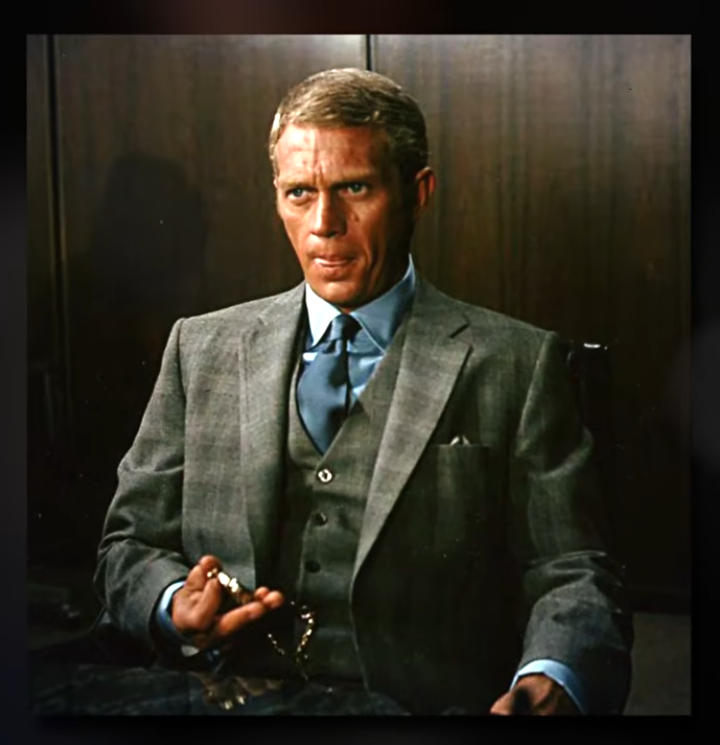 Steve McQueen seated in a grey three-piece suit and blue tie, holding a gold pocket watch in his right hand, looking directly at the camera