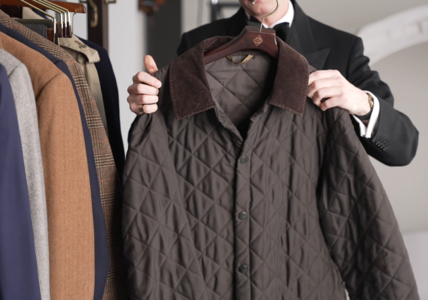Barbour Quilted Jacket Olive Brown Corduroy Collar Jack Collins holding up an olive-brown diamond-quilted Barbour jacket with a brown corduroy collar and snap front, with the wardrobe's suits and trench coat on the rail beside it