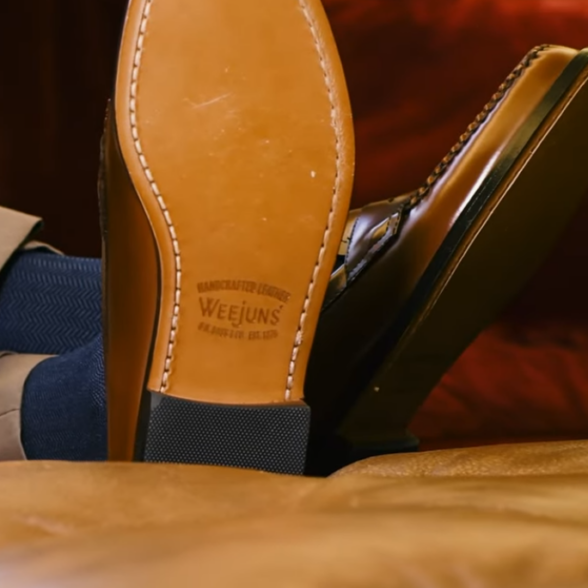 Close-up of the tan leather sole of a penny loafer with the Weejuns brand stamp, worn with navy socks and khaki trousers, feet crossed and resting on a leather surface