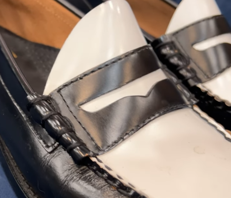Close-up of black and white spectator penny loafers showing the beefroll detail, where the leather is pinched into thick rolls at each end of the saddle strap
