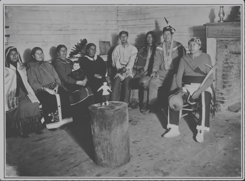 Historical black and white photograph of a Haudenosaunee (Iroquois) group of men, women, and a child seated indoors in traditional clothing, several wearing beaded moccasins