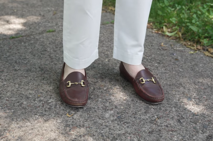 Brown leather horsebit loafers with gold hardware worn sockless with white trousers, standing on a gravel path