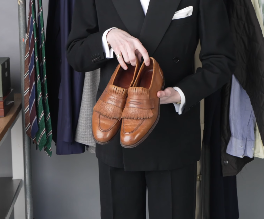 Light Brown Kiltie Loafers Slip On Jack Collins holding a pair of light brown leather kiltie loafers, with ties and suits on the rail behind him