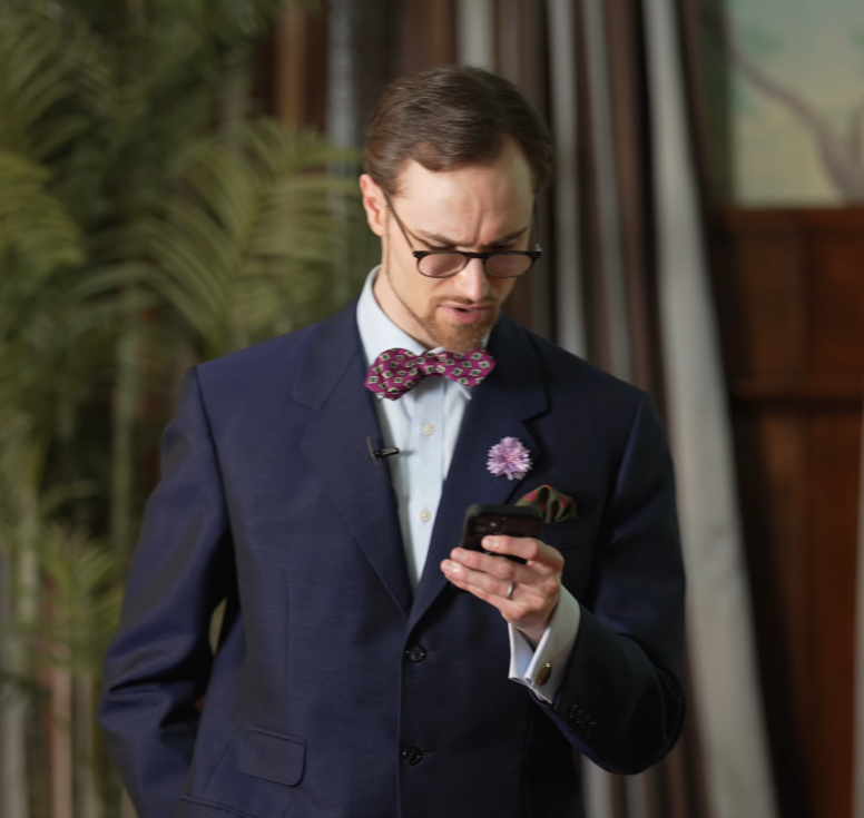 man reading heated comment thread phone A man in a navy suit with a patterned bow tie, boutonnière, and pocket square frowning at his phone