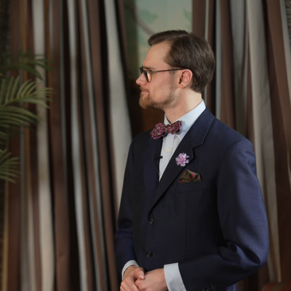 man voicing strong opinion navy suit bow tie A man in a navy suit with a patterned bow tie, boutonnière, and pocket square speaking with a serious expression, turned slightly away from the camera