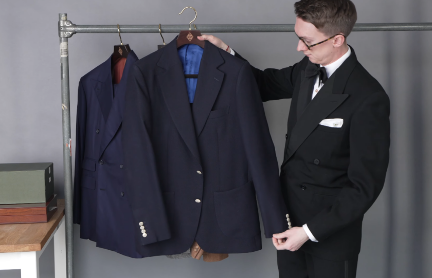Navy Hopsack Blazer Brass Buttons Half Lined Jack Collins holding up a navy hopsack blazer with brass buttons and patch pockets, showing the blue half-lining, with a navy double-breasted suit on the rail beside it