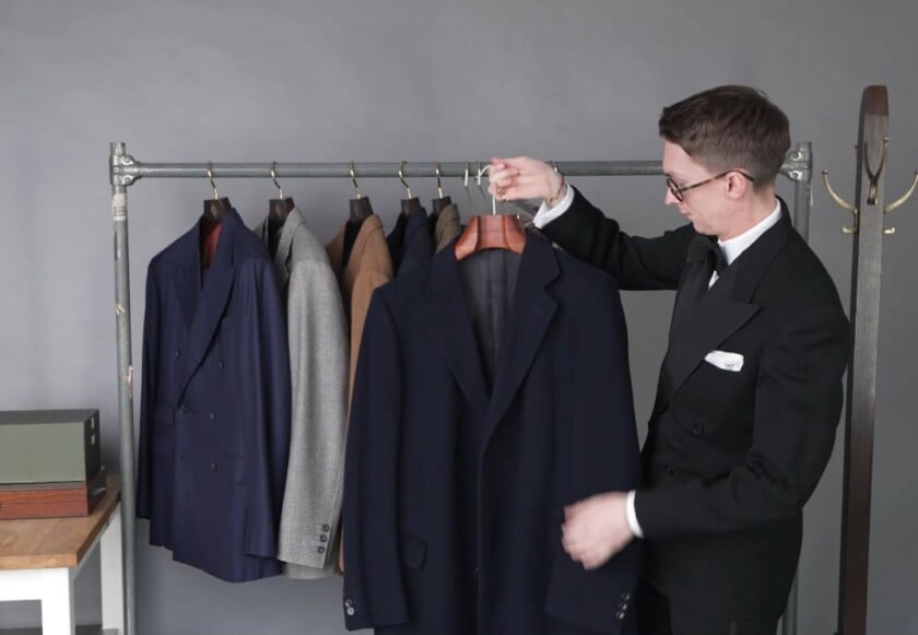 Navy Single Breasted Wool Overcoat Full Length Jack Collins hanging a navy single-breasted wool overcoat on a clothing rail, with the navy, gray, and brown suits from the wardrobe hanging behind it