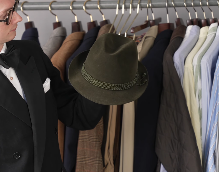 Olive Green Short Brim Trilby Braided Band An olive green short-brimmed Trilby with a braided band and feather detail, held in front of a clothing rail