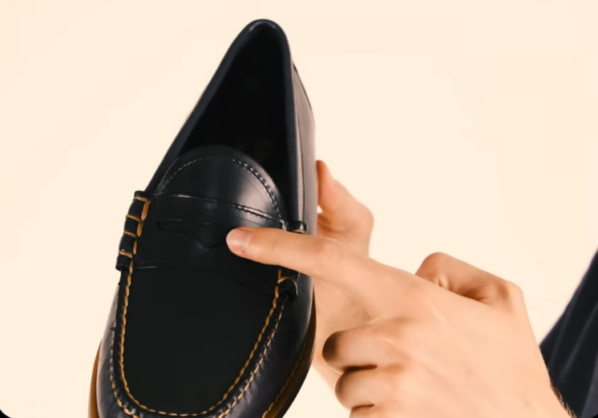 Close-up of a hand pointing to the diamond-shaped slot in the saddle strap of a black leather penny loafer, held against a cream background