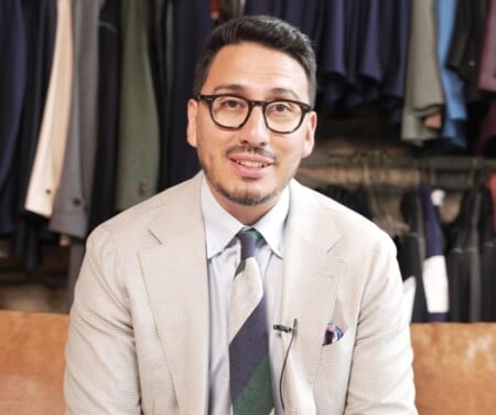 Custom clothier Ivan wearing a cream nailhead jacket, glasses, and a navy and green tie, seated against a wall of hanging jackets