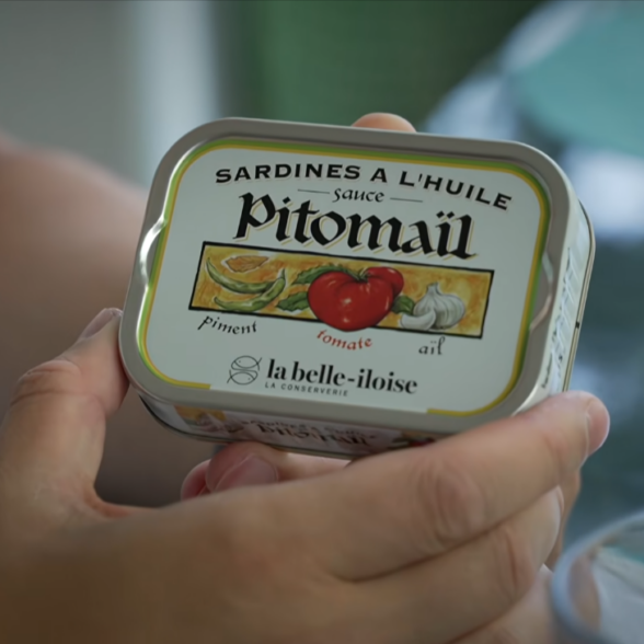 A hand holding a tin of La Belle-Iloise sardines in oil with Pitomaïl sauce
