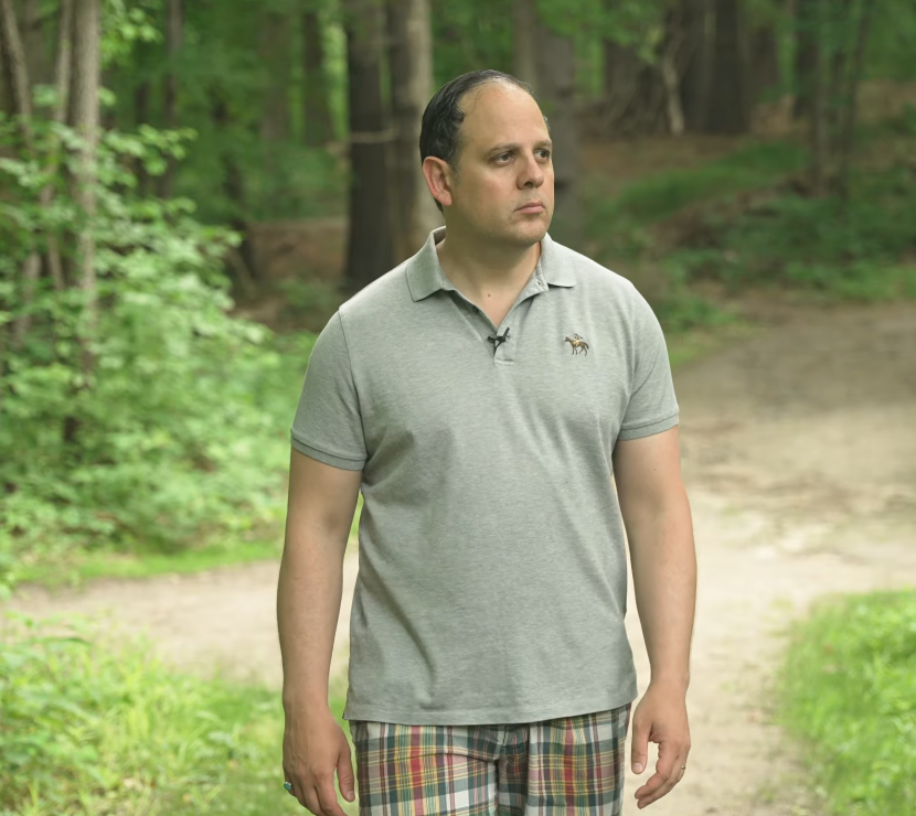 Pique Gray Polo Shirt Man wearing a heather gray piqué polo shirt with plaid shorts on a wooded path